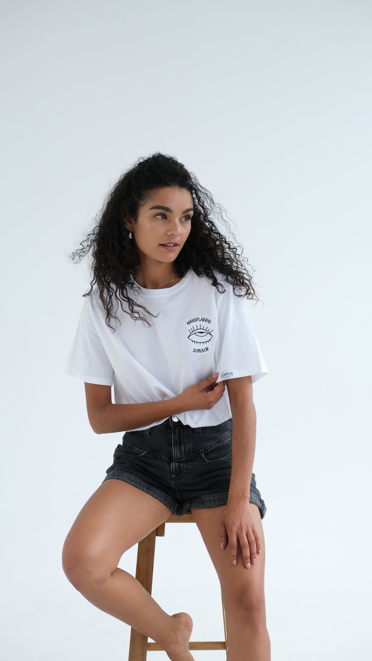 Woman perched on a stool wearing the white Mansplaining Survivor tee with denim shorts