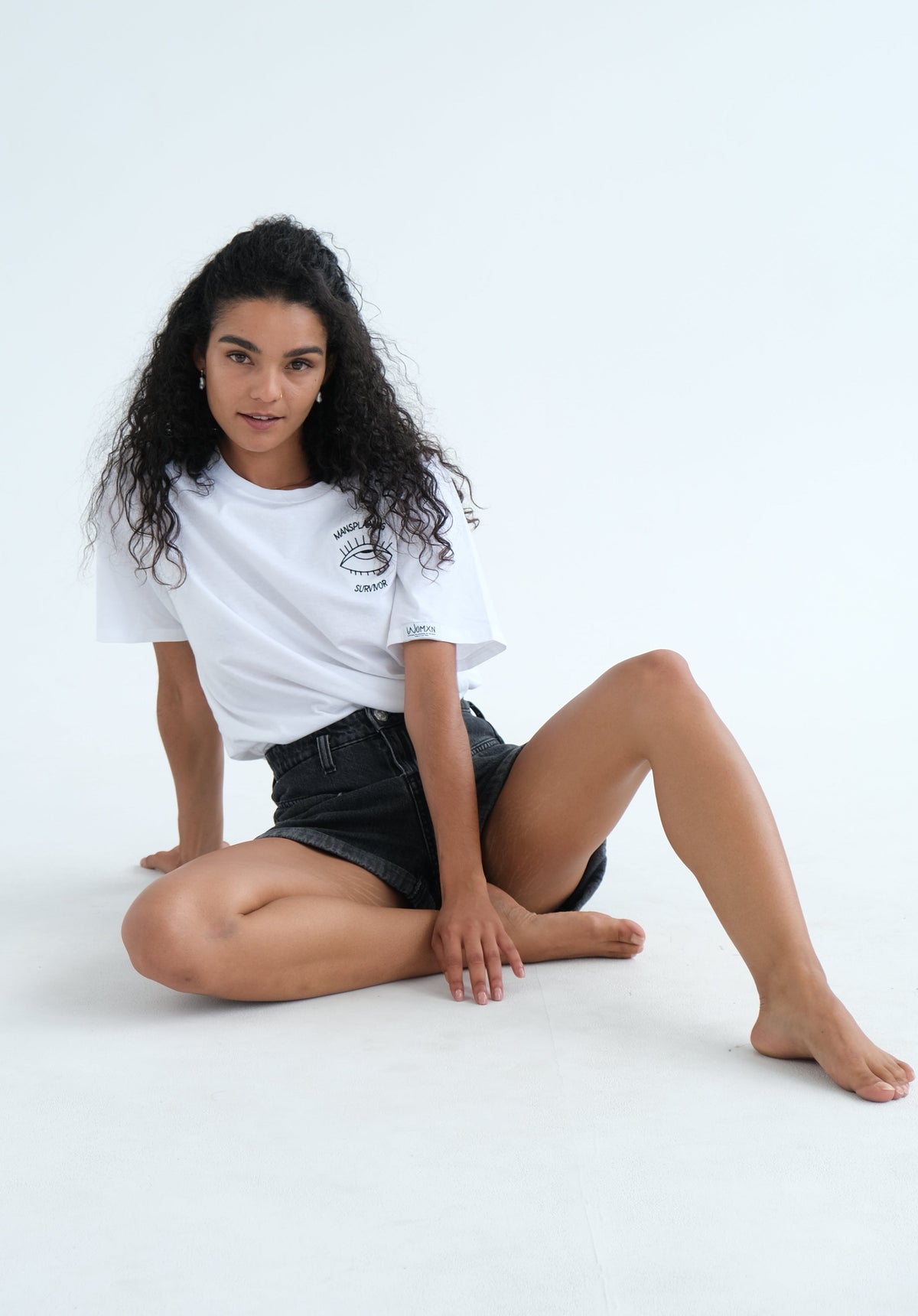 Woman sitting on the floor in the white Mansplaining Survivor tee and denim shorts