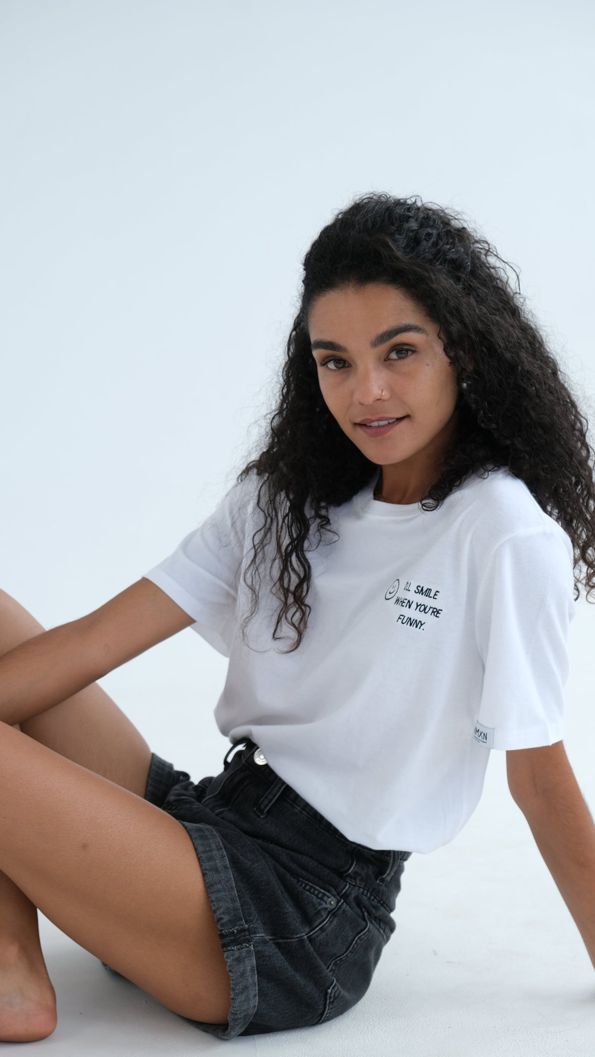 Woman sitting on the floor in the white I'll Smile When You're Funny tee and denim shorts