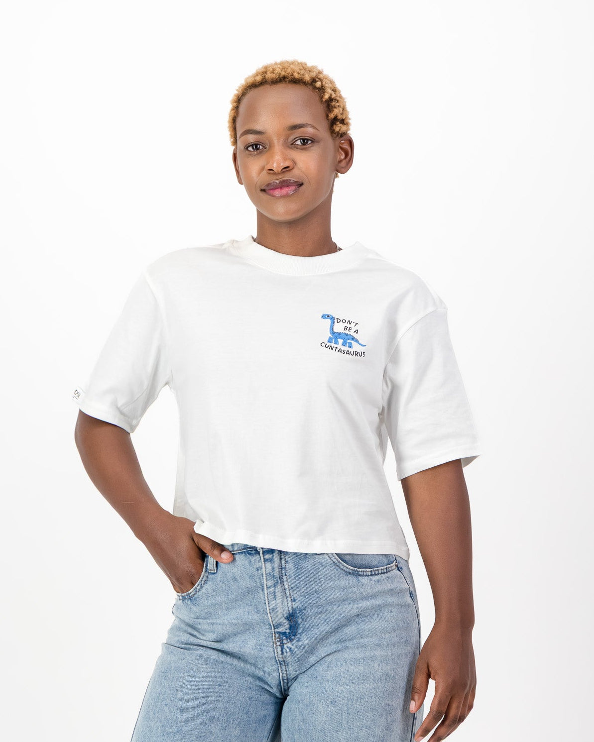 Woman wearing the white Don't Be A Cuntasaurus tee with its blue dinosaur embroidery and light blue jeans, front view