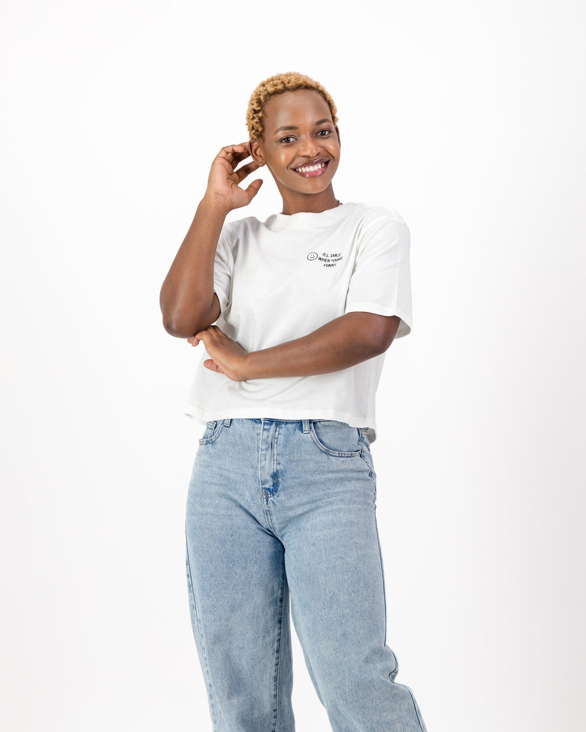 Smiling woman with one hand raised to her face in the white I'll Smile When You're Funny tee and jeans