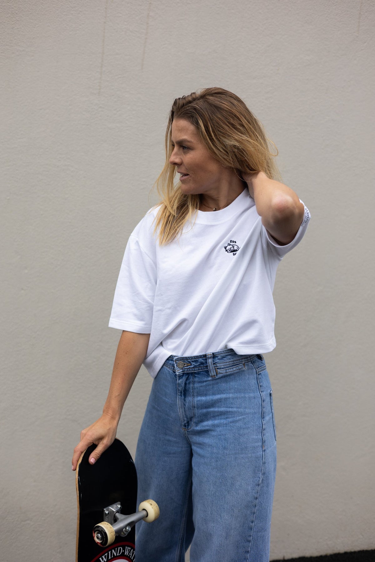 Woman with a skateboard and one hand behind her head in the white Cat Naps tee and jeans