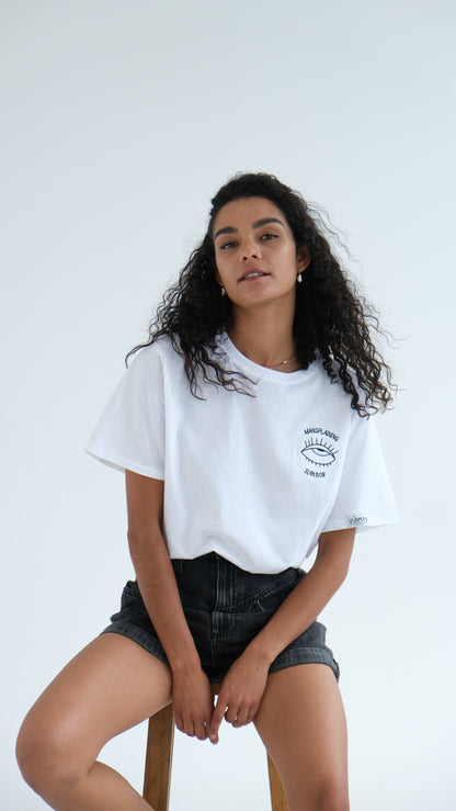 Woman sitting on a wooden stool in the white Mansplaining Survivor tee and black denim shorts