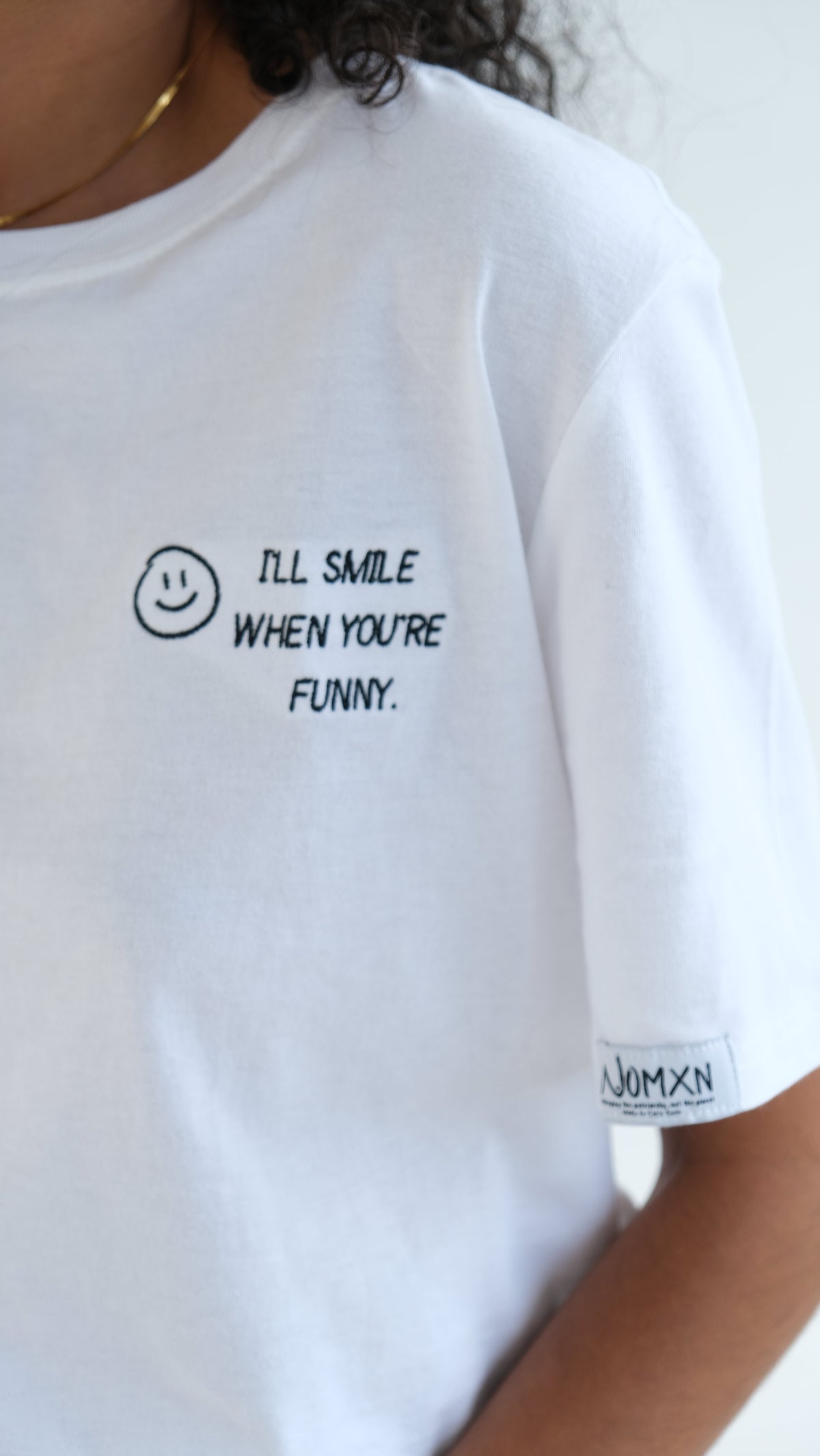 Close up of the smiley face embroidery and WOMXN sleeve label on the white I'll Smile When You're Funny tee