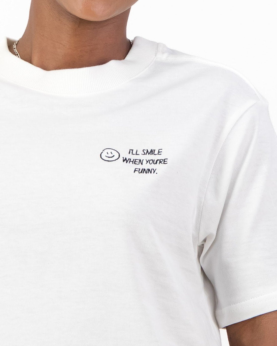 Close up of the black smiley face and I'll Smile When You're Funny embroidery on the white tee