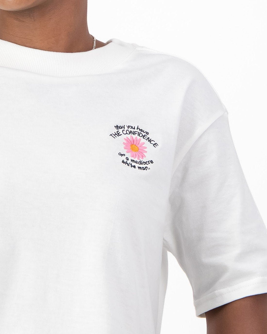 Close up of the pink daisy embroidery and May You Have the Confidence of a Mediocre White Man slogan on the white tee