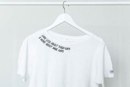 Close up of the Care Less About Thigh Gaps and More About Wage Gaps slogan along the collar of the white tee on a hanger
