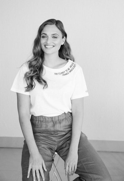 Black and white photo of a smiling woman sitting in the Too Clumsy to be Around Fragile Masculinity tee and wide trousers